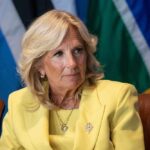 Jill Biden in a yellow blazer sitting, colorful flags behind.