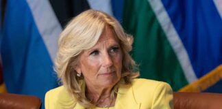 Jill Biden in a yellow blazer sitting, colorful flags behind.