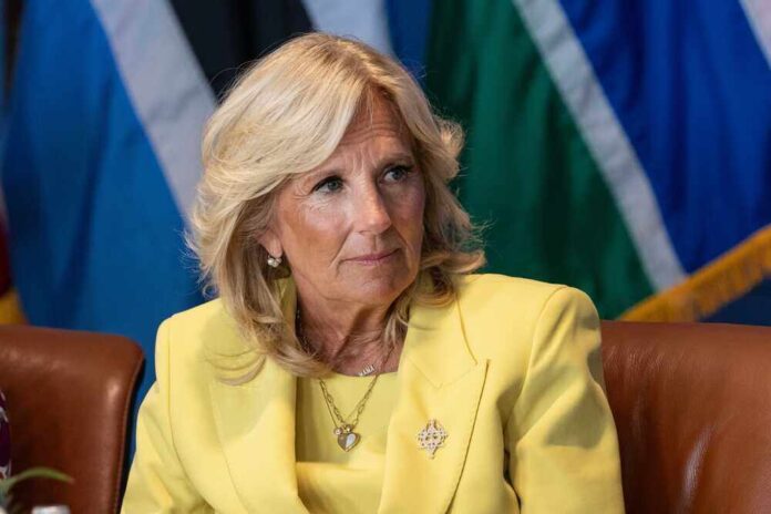 Jill Biden in a yellow blazer sitting, colorful flags behind.