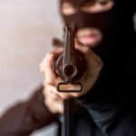Person in balaclava aiming rifle at the camera.