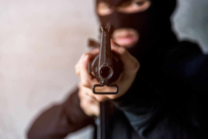 Person in balaclava aiming rifle at the camera.