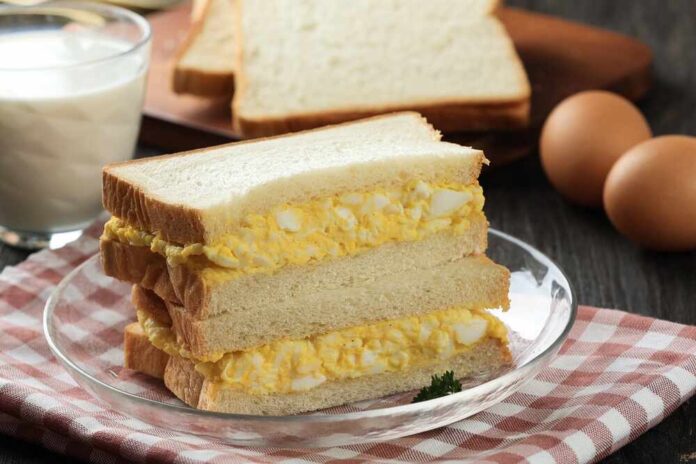 shutterstock_2663394607.jpg Three-layered egg sandwich on a plate with a glass of milk and eggs in the background