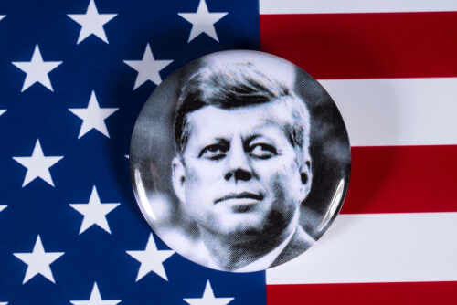 A black and white campaign button featuring a portrait of John F. Kennedy on an American flag background