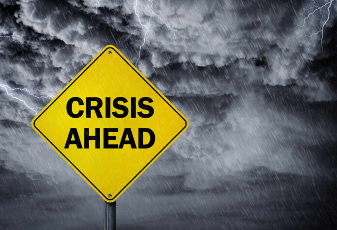 shutterstock_576862294.jpg A yellow warning sign that reads 'CRISIS AHEAD' against a stormy sky