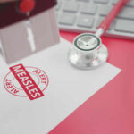 A stamp marking 'MEASLES ALERT' on a piece of paper next to a stethoscope and keyboard