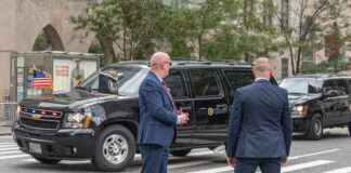 Two security personnel monitoring a convoy of black SUVs in an urban area