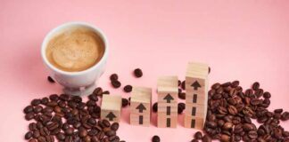 A cup of coffee next to wooden blocks with upward arrows and scattered coffee beans on a pink background