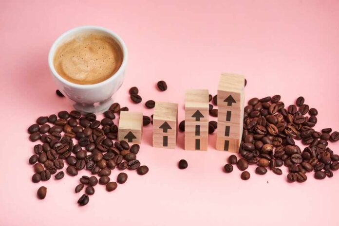 A cup of coffee next to wooden blocks with upward arrows and scattered coffee beans on a pink background