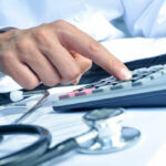 Americans Worried as Healthcare Becomes Top Cost Fear Doctor using calculator with stethoscope on the table.