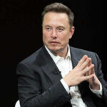 Man in suit, hands clasped, black background.