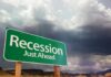 A green road sign indicating 'Recession Just Ahead' against a cloudy sky