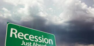 A green road sign indicating 'Recession Just Ahead' against a cloudy sky