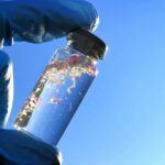 A gloved hand holding a glass vial containing colorful microplastics against a blue sky