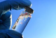 A gloved hand holding a glass vial containing colorful microplastics against a blue sky