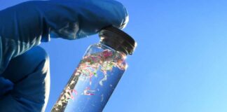 A gloved hand holding a glass vial containing colorful microplastics against a blue sky