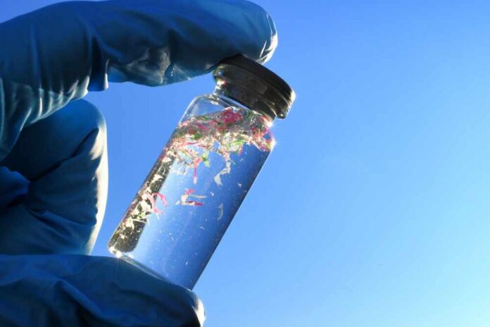 A gloved hand holding a glass vial containing colorful microplastics against a blue sky