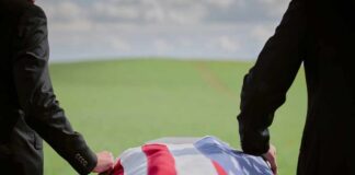 Two individuals standing beside a flag-draped casket in a green field