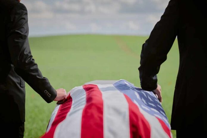 shutterstock_2665284487.jpg Two individuals standing beside a flag-draped casket in a green field