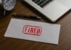 Another 8,000 Jobs Cut — AI’s Costly Reality Check An envelope marked 'FIRED' on a wooden desk next to a clock and a laptop