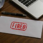 Another 8,000 Jobs Cut — AI’s Costly Reality Check An envelope marked 'FIRED' on a wooden desk next to a clock and a laptop