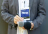 Hit Machine Silenced — Power Vacuum Looms Journalist with press badge, notebook, and microphones.
