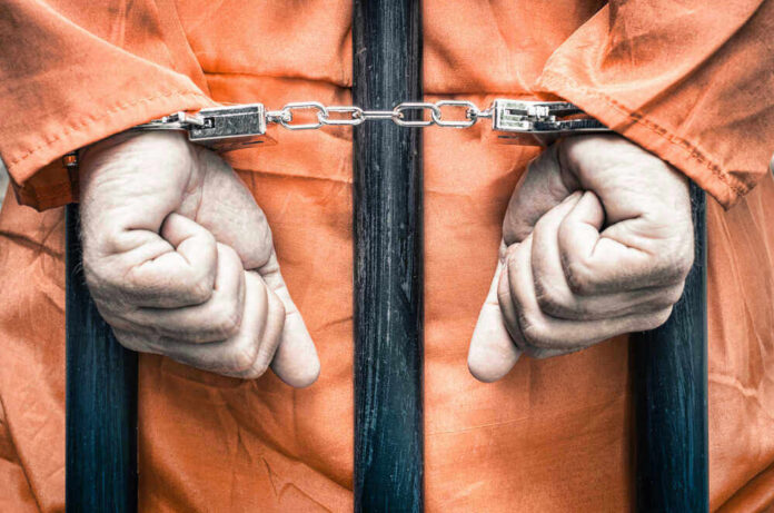 shutterstock_217094626 Handcuffed person in orange jumpsuit behind prison bars.