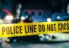 Grim Stench Leads To School Horror Police line caution tape at a crime scene with blurred figures in the background