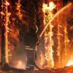 Helicopter fighting massive forest fire with water bucket.