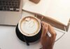 A hand holding a coffee cup with latte art next to a laptop and an open notebook