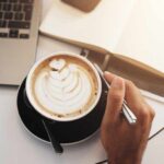 Coffee Shock: Heart Risk Drops A hand holding a coffee cup with latte art next to a laptop and an open notebook