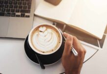 A hand holding a coffee cup with latte art next to a laptop and an open notebook