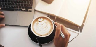 A hand holding a coffee cup with latte art next to a laptop and an open notebook