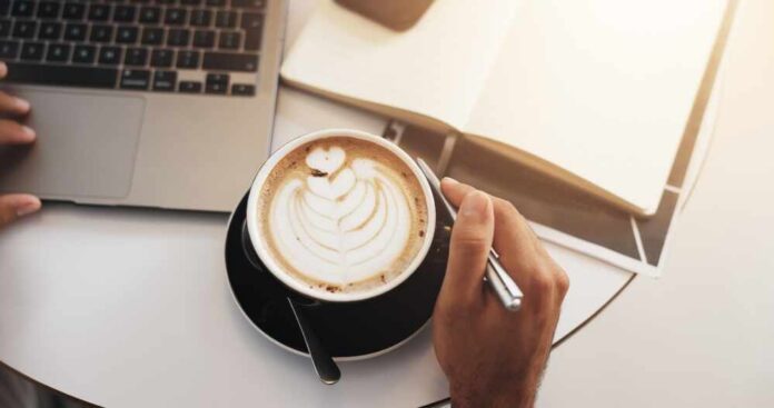 Coffee,Shop,,Laptop,And,Above,Of,Hands,With,Notebook,For A hand holding a coffee cup with latte art next to a laptop and an open notebook