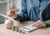 Billionaire Cash Bomb Hits Kids’ Accounts Person placing a coin into a piggy bank while using a calculator