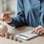 Billionaire Cash Bomb Hits Kids’ Accounts Person placing a coin into a piggy bank while using a calculator