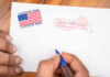 Ballot Gatekeeping? USPS Demands Names, Barcodes Hands addressing an official election mail envelope