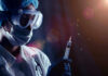 Person in PPE holding syringe with liquid droplets.