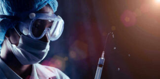 Person in PPE holding syringe with liquid droplets.