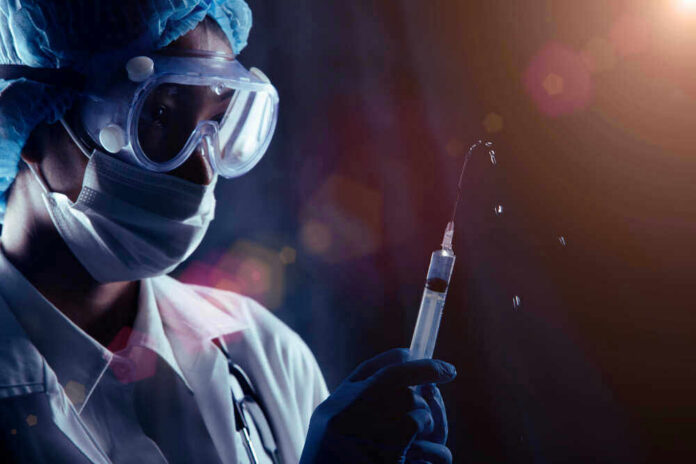 shutterstock_1880299123 Person in PPE holding syringe with liquid droplets.