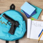Grandfather’s Gun, School Carnage A blue backpack with a handgun and school supplies on a wooden surface