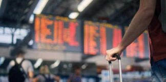 A traveler holding a suitcase in an airport terminal with a flight information display in the background