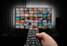 TV Icon Exits: Will Vanish In October Hand holding a remote control in front of a television displaying various media options