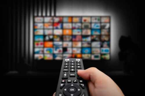 Vod,Service,Screen.,Man,Watching,Tv,With,Remote,Control,In Hand holding a remote control in front of a television displaying various media options
