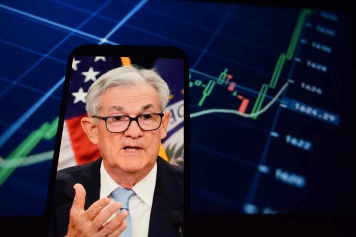 shutterstock_2615221169 Jerome Powell speaking on a smartphone screen with stock chart in background