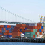 A view of stacked shipping containers at a port with a bridge in the background