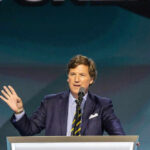 Tucker SLAMMED Over Hitler Spin Tucker Carlson speaking at a podium with a microphone