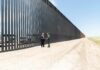 Judge Slams Brakes On Border Wall Two people stand by a tall border fence on a dusty road