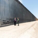 Judge Slams Brakes On Border Wall Two people stand by a tall border fence on a dusty road