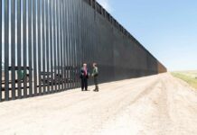 Two people stand by a tall border fence on a dusty road