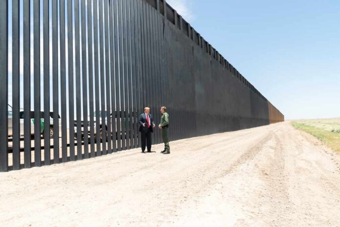 Two people stand by a tall border fence on a dusty road